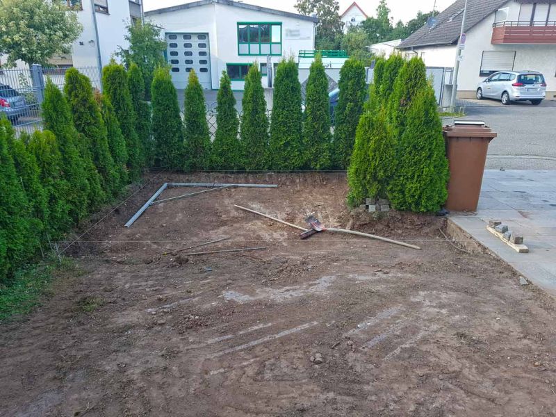 Eine vorbereitete Erdfläche, umgeben von hohen Lebensbäumen. Werkzeuge und Begrenzungen liegen bereit. Im Hintergrund eine Straße mit geparkten Autos.