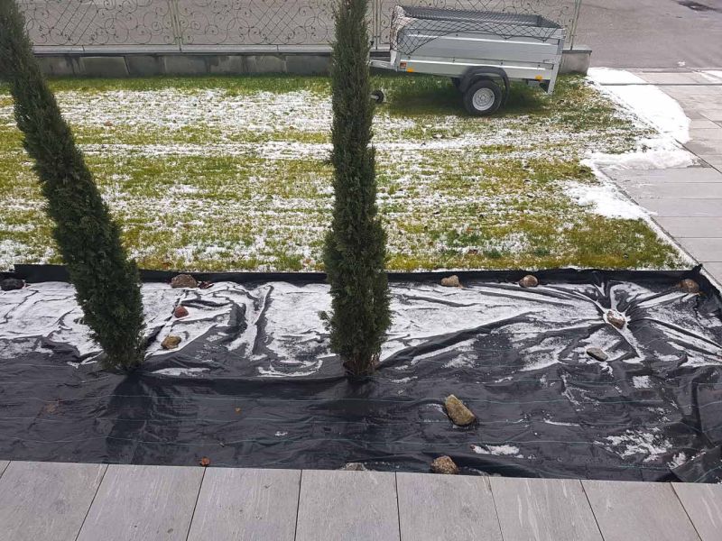 Ein Parkplatz mit Rasengittern, teils von Schnee bedeckt. Vlies mit Steinen bedeckt Pflanzbereich. Anhänger und Metallzaun im Hintergrund.