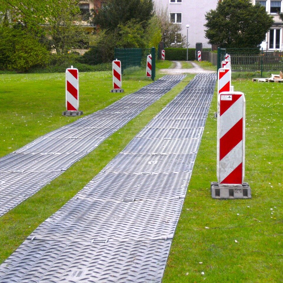 Bodenschutzplatten bilden eine Fahrstraße über eine Rasenfläche, gesichert mit Warnbaken für sichere Zufahrt.