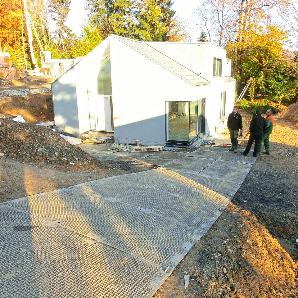 Temporäre Fahrstraße aus Bodenschutzplatten führt zur Baustelle eines modernen Wohnhauses auf unebenem Gelände.