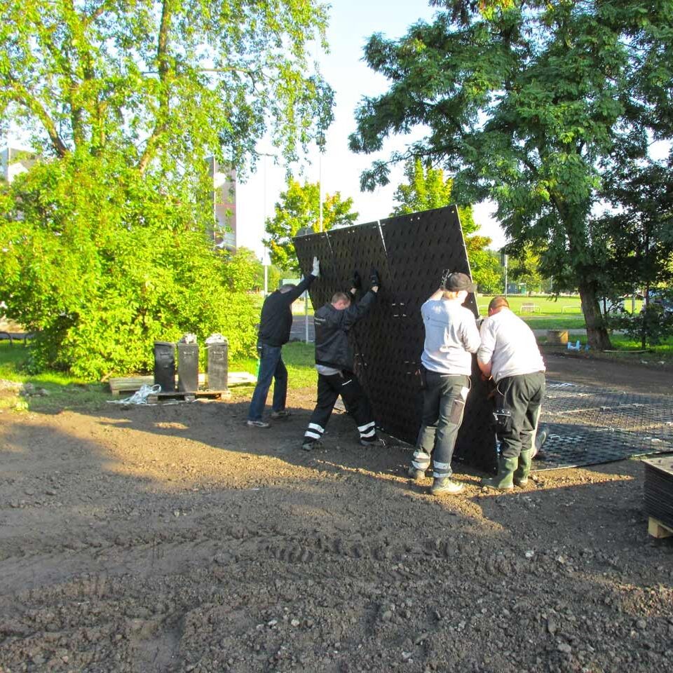 Mehrere Arbeiter heben große Bodenschutzplatten an, um sie auf einer Baustelle zu verlegen.