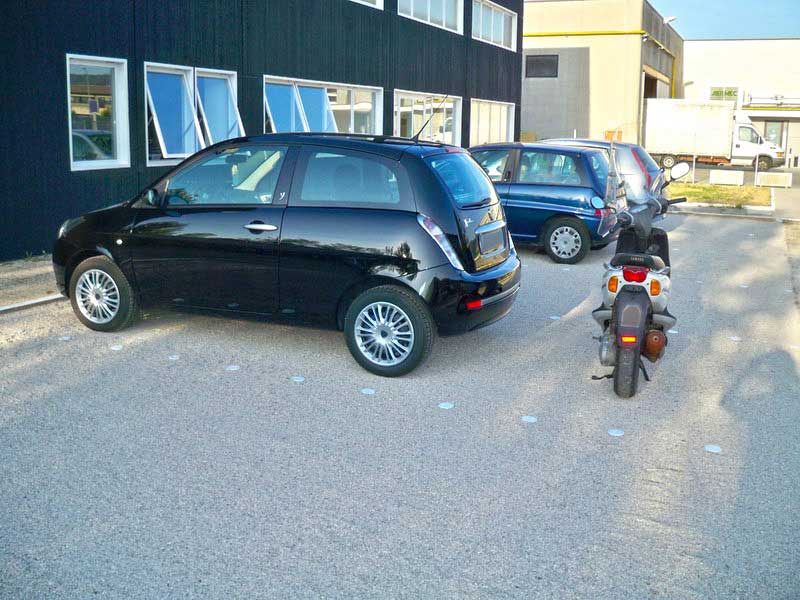 Fahrzeuge parken auf einer stabilen, hellen Kiesfläche vor einem Bürogebäude.